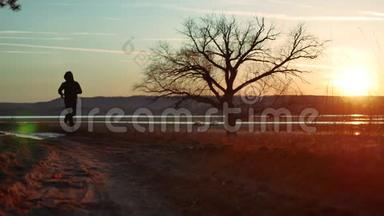 剪影人在日落时沿着道路奔跑，独自站在树上剪影。 在大自然中奔跑的运动青年